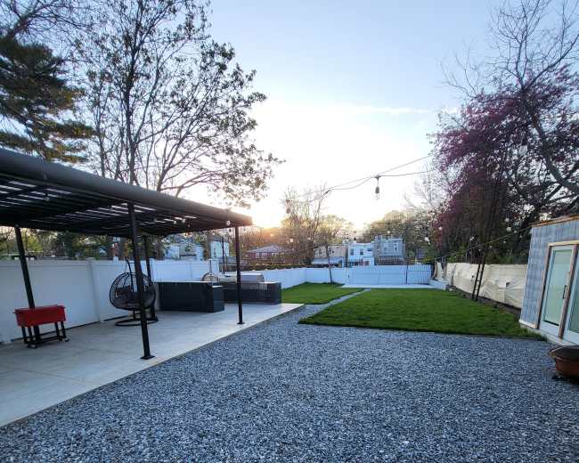 The image shows a gravel-covered backyard with a patio area, a green lawn, and a structure equipped with a roof.