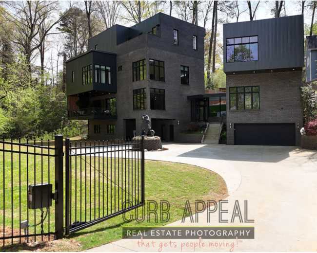 The image shows a modern, multi-level home with a dark facade, large windows, and a circular driveway, set within a wooded area.