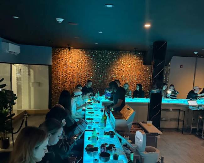 A bar area with patrons sitting at a glowing blue countertop, engaging with their phones while various items are placed on the surface in front of them.