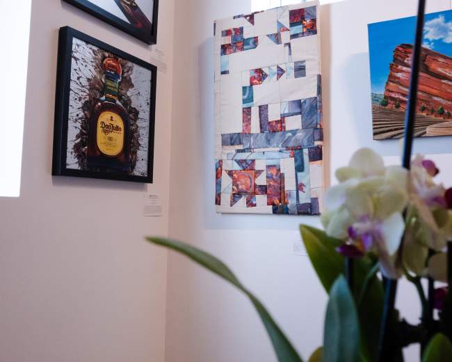 The image shows a gallery wall featuring three distinct pieces of framed artwork, including a bottle-shaped painting and a mixed media canvas, alongside a vase of orchids on a table in the foreground.