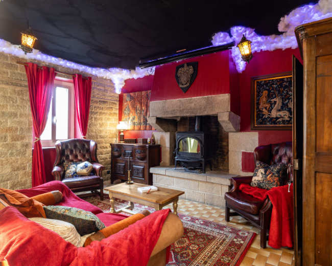 The image shows a cozy living room with a stone fireplace, red walls, and traditional furniture, including leather armchairs and a wooden cabinet.
