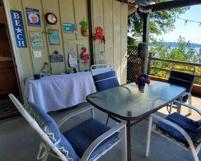 The image shows a covered patio area with a dining table and chairs overlooking a body of water, decorated with beach-themed signs and plants.