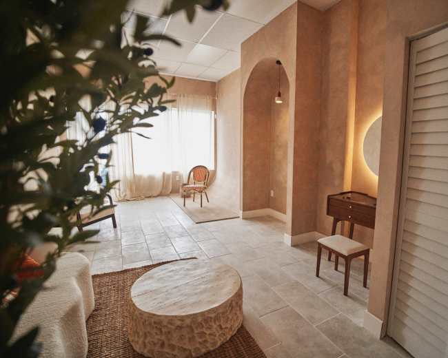 The image shows a softly lit interior space with neutral-colored walls, a round stone coffee table, a minimalist wooden chair, and a small dresser beneath a circular mirror.