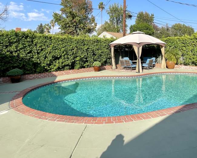 The image shows a residential backyard featuring a swimming pool surrounded by a concrete deck and a shaded seating area under a gazebo.
