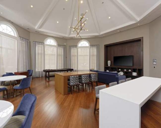 A spacious room with a high ceiling, featuring a seating area with multiple tables and chairs, large windows, and a media console with a television.