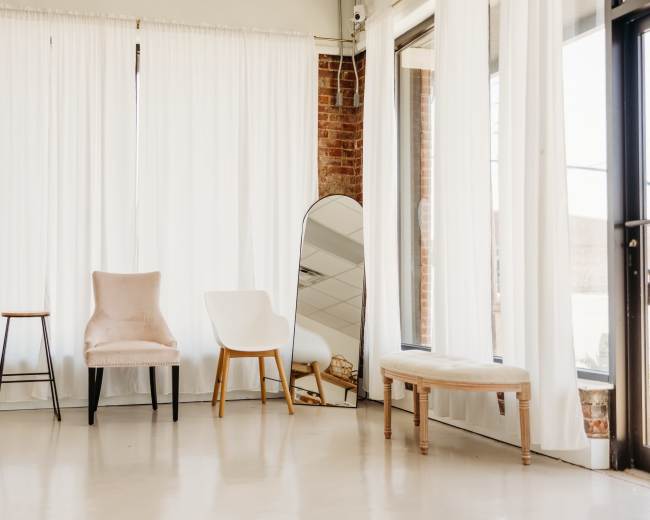 The scene shows a well-lit room with three chairs of different styles, a large mirror, and white curtains against a brick wall.