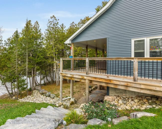 A blue house with a wooden deck overlooks a body of water, surrounded by trees and landscaped with stone steps and greenery.