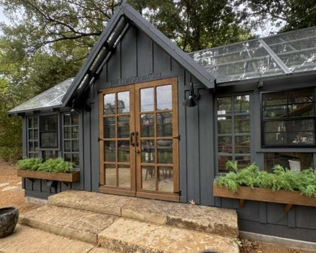 The image shows a modern gray garden conservatory with large glass windows and wooden doors, flanked by planters filled with greenery, set against a backdrop of trees.