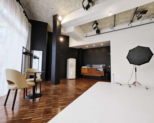 A modern photography studio features a white backdrop, a kitchenette with a refrigerator, and a table set with two chairs.