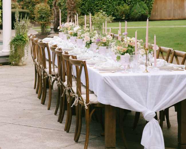 A long wooden dining table is set outdoors with white tablecloths, pink candles, and floral centerpieces, surrounded by lush greenery and string lights overhead.