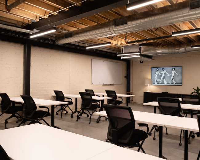 The image shows an empty training room with multiple rows of white tables and black chairs, featuring a wall-mounted screen displaying a monochrome image.