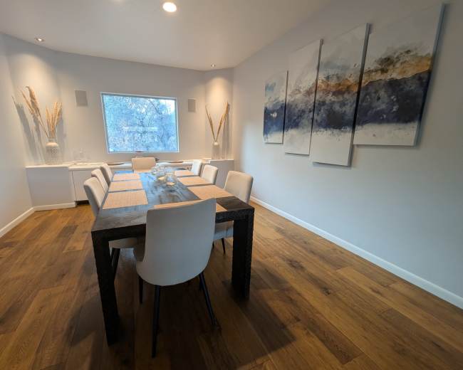 A modern dining room features a long table set with placemats, surrounded by white chairs, under a window with a view of trees.