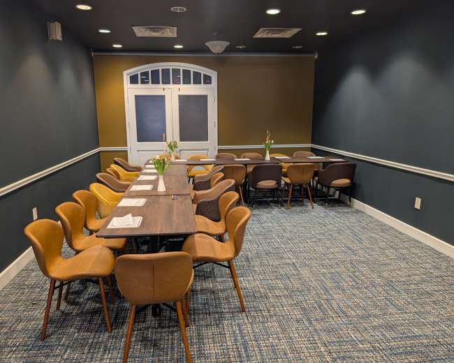 A long wooden table with chairs surrounds it in a dimly lit meeting room featuring a patterned carpet and a pair of double doors at the far end.