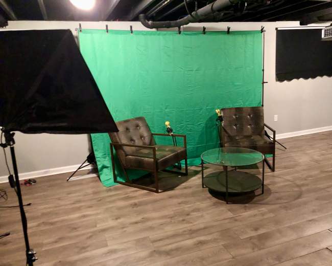 A studio setup with two chairs, a glass table, and a green backdrop, illuminated by softbox lighting.