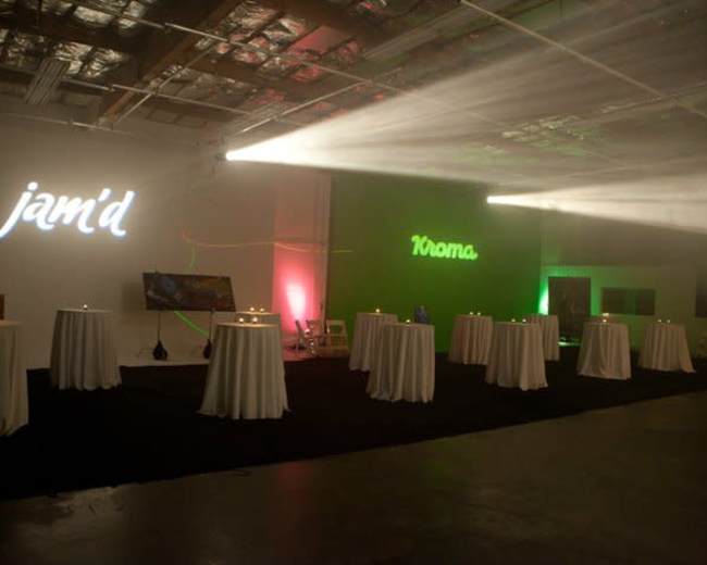 The image shows a spacious indoor venue with several round tables draped in white cloth, illuminated by colored lights, and featuring the words "jam'd" and "Kroma" projected on the walls.