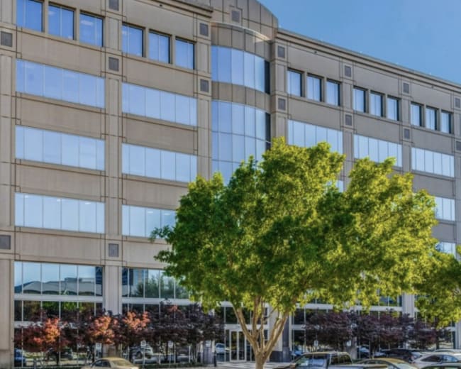 The image shows a modern office building with large glass windows and surrounding trees in an urban setting.