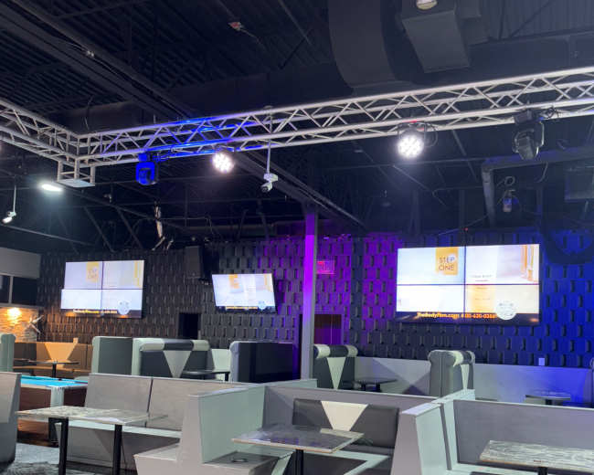 The image shows an interior of a modern lounge or nightclub featuring gray seating booths, multi-colored lighting, and large screens displaying information.