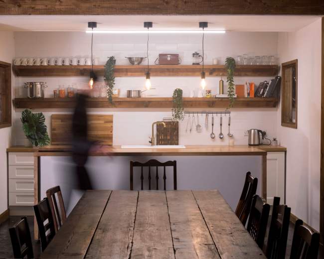 A wooden table sits in the foreground of a modern kitchen featuring open shelves with various kitchenware and plants, illuminated by pendant lights.