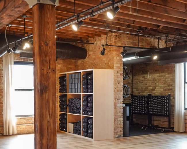 The image shows a large, open room with wooden floors, exposed brick walls, and gym equipment neatly arranged in the background.