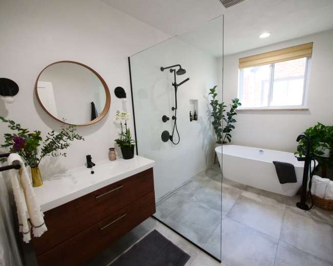 A modern bathroom features a glass shower with a rainfall showerhead, a freestanding bathtub, and a double sink vanity with a circular mirror and plants arranged decoratively.