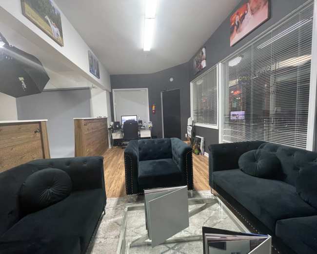The image shows a modern office waiting area featuring three black upholstered sofas around a glass coffee table, with large windows and gray walls.