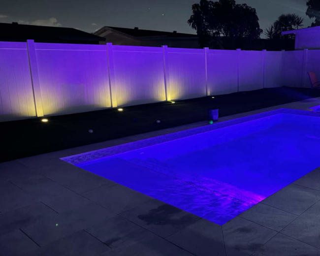 The image depicts a modern swimming pool illuminated with purple lights, surrounded by a white fence and gray tiled patio in a nighttime setting.