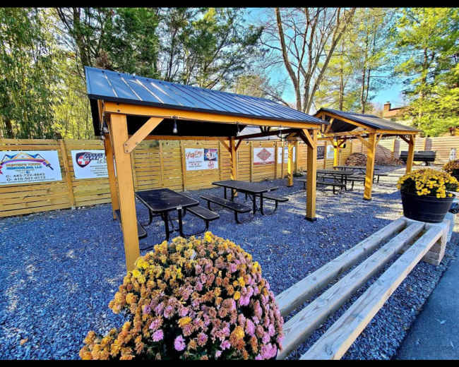 The image shows an outdoor seating area with wooden pavilions, picnic tables, and flower planters surrounded by wooden fencing.