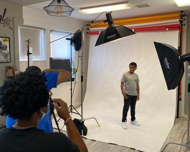 A young boy stands posing in front of a white backdrop, while a photographer adjusts equipment in a well-lit studio.