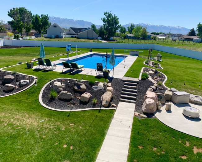 A backyard features a swimming pool surrounded by lounge chairs, landscaped stone paths, and a grassy area with a white fence in the background.