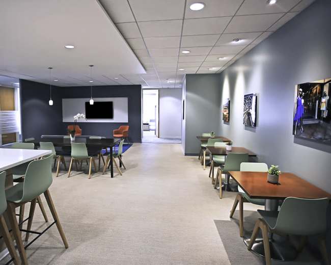 The image shows a modern office break room featuring tables with chairs, a large countertop, and a wall-mounted screen.