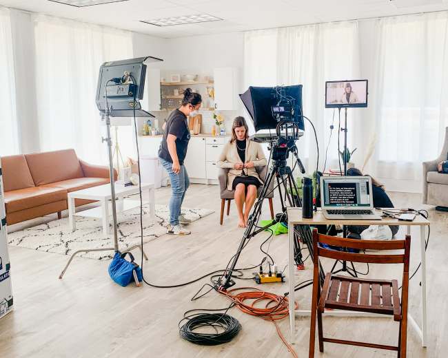A woman sits on a chair in a bright room set up for filming, with cameras and lights arranged around her.