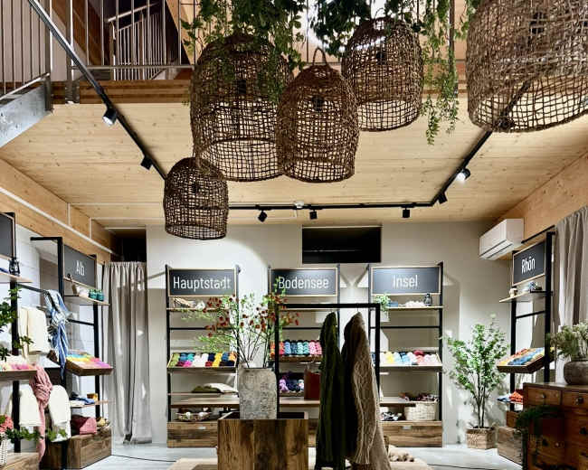 The image shows a retail space featuring wooden displays, clothing racks with colorful apparel, and pendant lights, all set against a minimalist interior design.