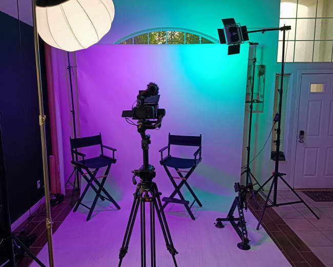 A professional photography setup with two director chairs, a camera on a tripod, and colorful lighting in a room with a front door visible in the background.