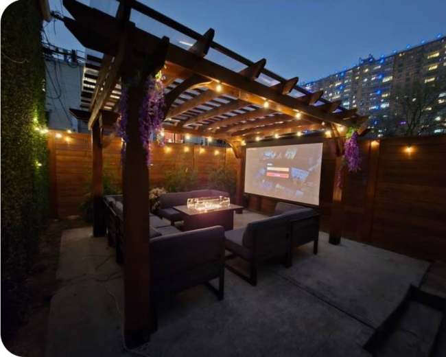 The image shows an outdoor patio with a wooden pergola, string lights, and a large screen displaying a movie or presentation, surrounded by urban buildings in the background.