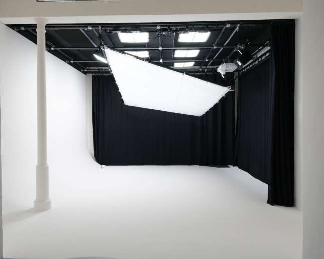 The image shows a professional photography studio with a white backdrop, black curtains, and overhead lighting fixtures.