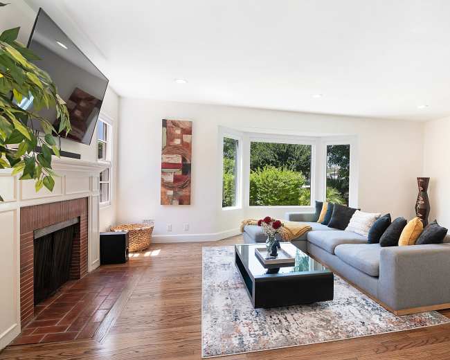 The living room features a gray sofa, a coffee table, and a large window with views of greenery, along with a flat-screen TV mounted on the wall.