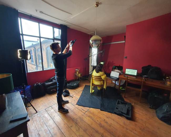 A person is setting up a camera to photograph a model sitting on a chair in a room with red walls and a disco ball hanging from the ceiling.