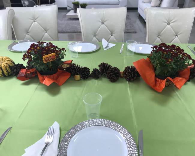 A dining table is set with white plates, utensils, and two flower arrangements, surrounded by decorative pinecones and gourds on a green tablecloth.