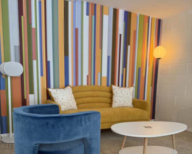 The image shows a modern living room featuring a yellow sofa with decorative pillows, a blue chair, and a white coffee table on a patterned carpet against a colorful striped wall.