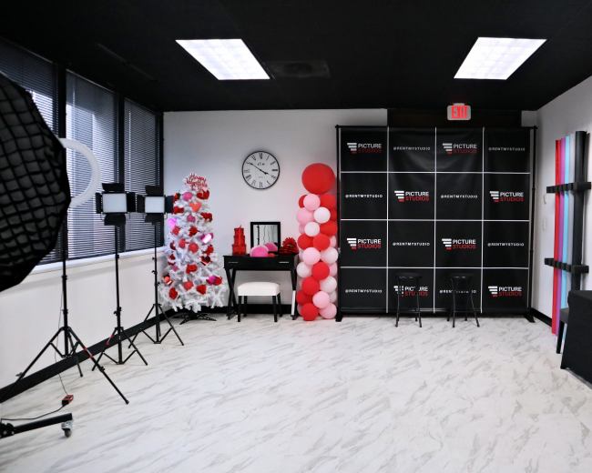 A photo studio setup featuring a decorative backdrop with black and white panels, a clock on the wall, lighting equipment, and a display of balloons in red and pink hues.