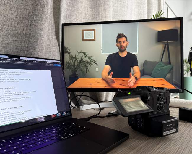 A laptop shows a text document next to a camera and a monitor displaying a person speaking in front of a wooden table.