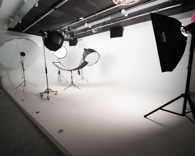 A photography studio with a plain white backdrop, lighting equipment, and a curved chair set up in the center.