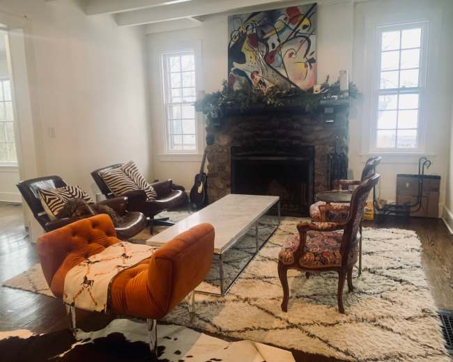 The room features a stone fireplace with a mantel adorned with decorations, surrounded by various seating options, including two armchairs, a modern couch, and a coffee table on a patterned rug.