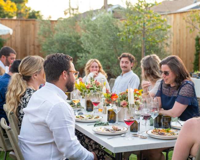 A group of people enjoys a dinner gathering outdoors at a table adorned with floral centerpieces and wine glasses.