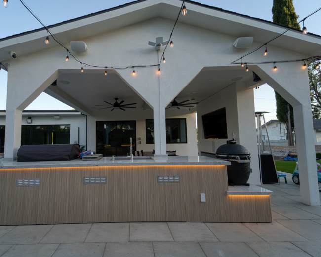 The image shows a modern patio area with a large overhang, string lights, a barbecue grill, and a seating space.