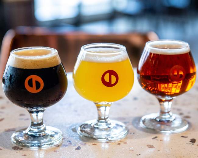 Three distinct glasses of beer are placed on a speckled countertop, featuring a dark brew, a golden lager, and a reddish amber beer, each with a circular logo on the side.
