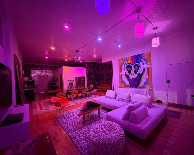 The image shows a modern living space with a white sectional sofa, colorful lighting, an art piece on the wall, and a dining area visible in the background.