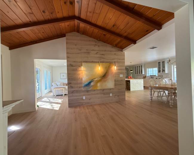 The image shows an interior space featuring a wood-beamed ceiling, a wooden accent wall with a painting, and an open layout connecting to a kitchen area in the background.