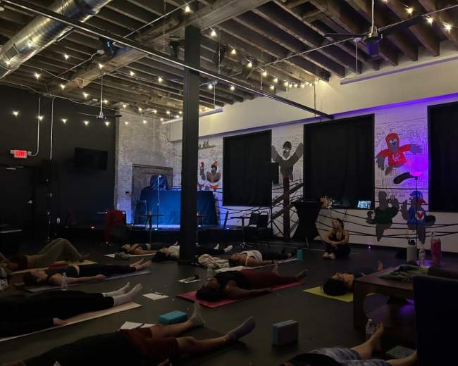 A group of people practice yoga on mats inside a dimly lit studio featuring exposed beams and string lights, while a mural adorns one wall.
