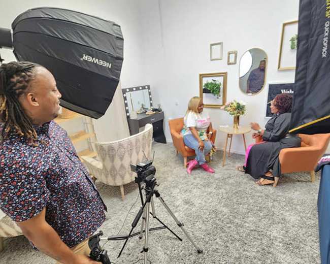 A man stands with a camera on a tripod while two women sit in an indoor setting decorated with plants and framed artwork.